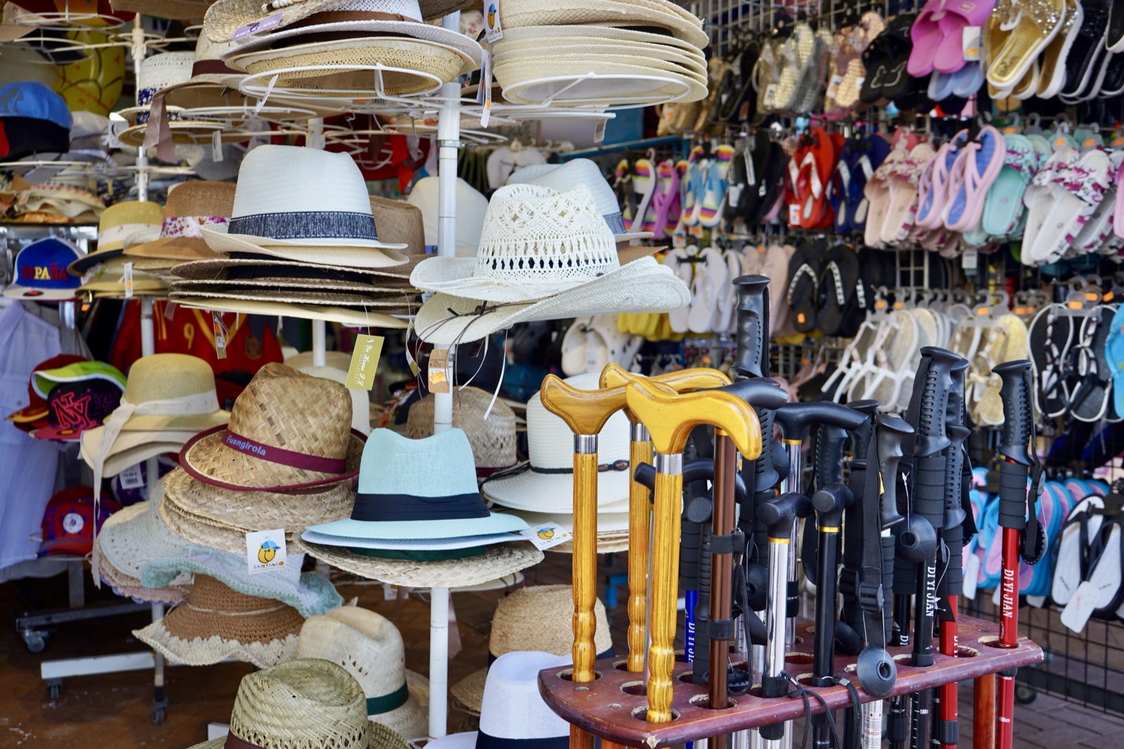 Market stall with straw hats, flip-flops, and a rack where walking canes stand mixed with trekking poles