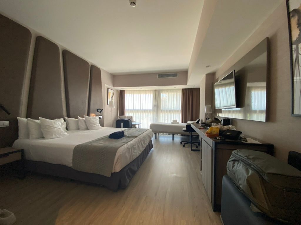 A spacious hotel room with a wide bed, a second bed by the window, a desk with a chair and a wall-mounted TV, and open floor space between them.
