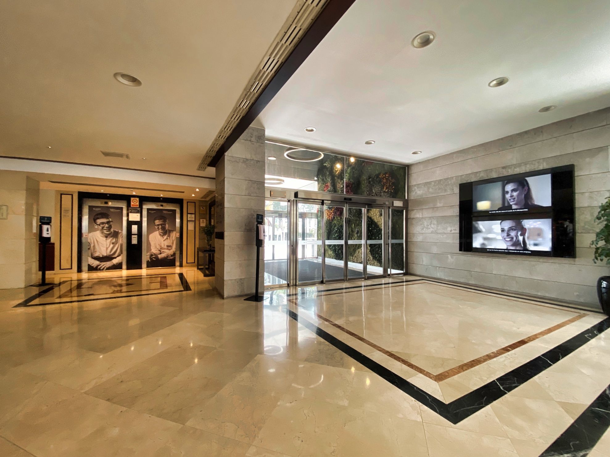 The entrance hall of ILUNION Malaga seen from inside. Sliding glass doors in the middle, two lifts to the left with large portraits of smiling staff members on their doors, and a screen on the right wall showing a video of a woman speaking.