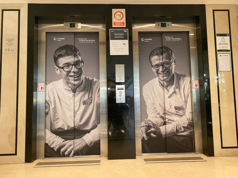 Two lift doors side by side, each carrying a large black-and-white portrait of a smiling staff member in a white uniform, with the text "Hoteles con Todos Incluidos" above.