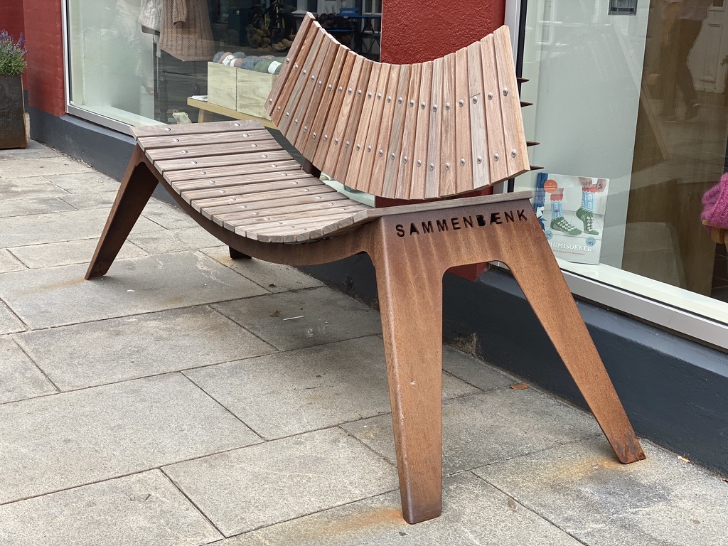 A bench on a pavement outside a shop window. Its wooden seat slopes inward from both ends and the backrest curves, so people sitting on it are turned towards each other. The word Sammenbænk is cut into the rusted steel frame.