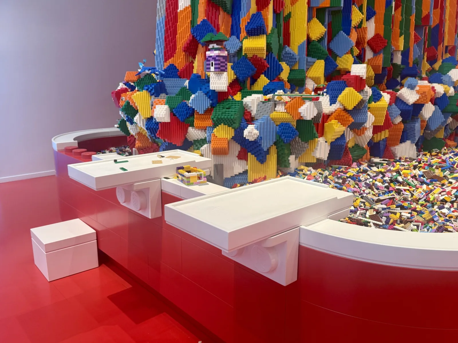 A curved red counter holds a pool of loose LEGO bricks below a tall structure built of bricks in every colour. Two white trays stick out from the side of the counter, held by brackets shaped like large LEGO bricks, with a few bricks on the left one. A loose white box stands on the red floor to the left.