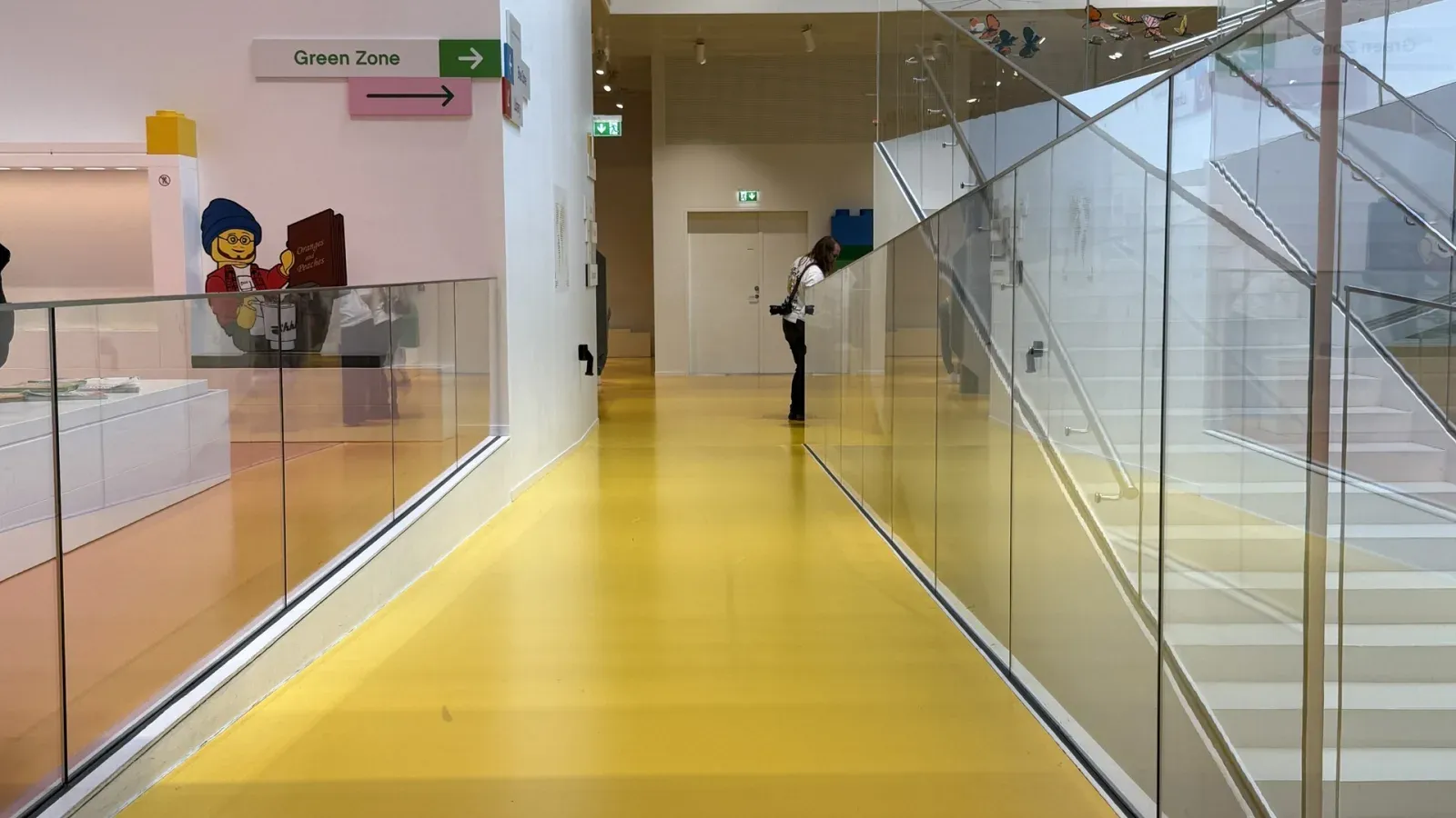 A wide yellow floor ramps up toward the viewer inside LEGO House, with glass balustrades on both sides. Behind the glass on the right, a white staircase rises the other way. A sign on the wall reads Green Zone, with an arrow.