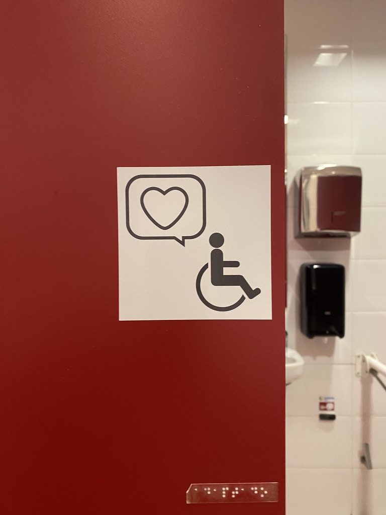 A dark red toilet door with a white sign showing a wheelchair symbol and a speech bubble with a heart in it. Below the sign sits a braille plate. Through the open door, a grab rail and a soap dispenser are visible.